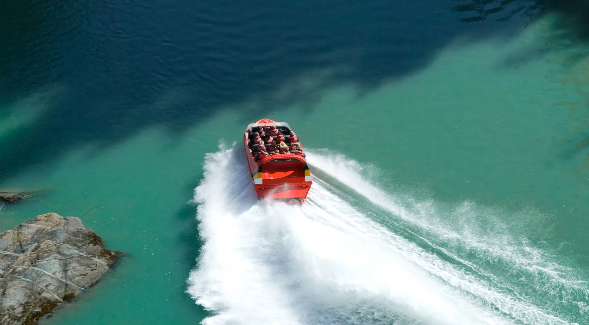 A red jet boat carving a white wake across clear turquoise water beside a rock outcrop