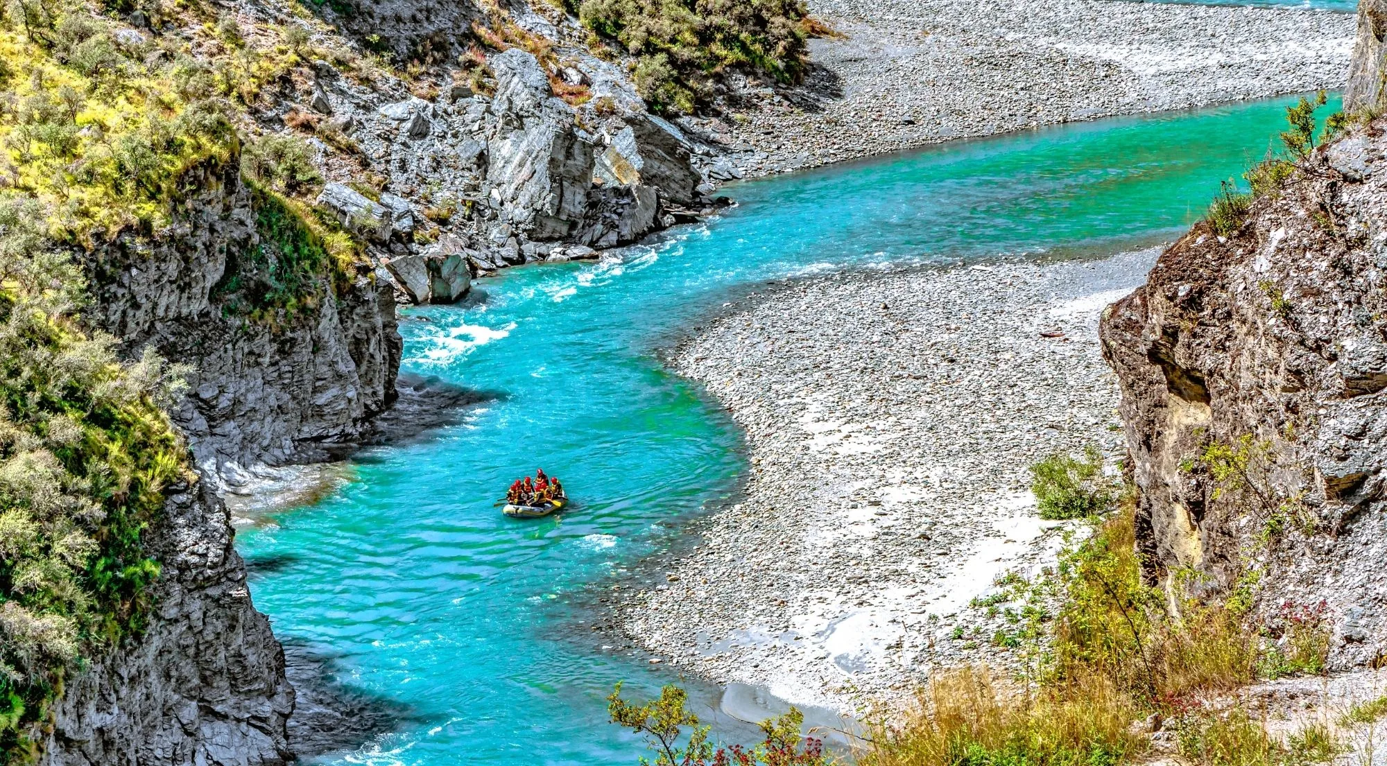 Paddlers in a red raft on bright turquoise water between a rock wall and a shingle bank