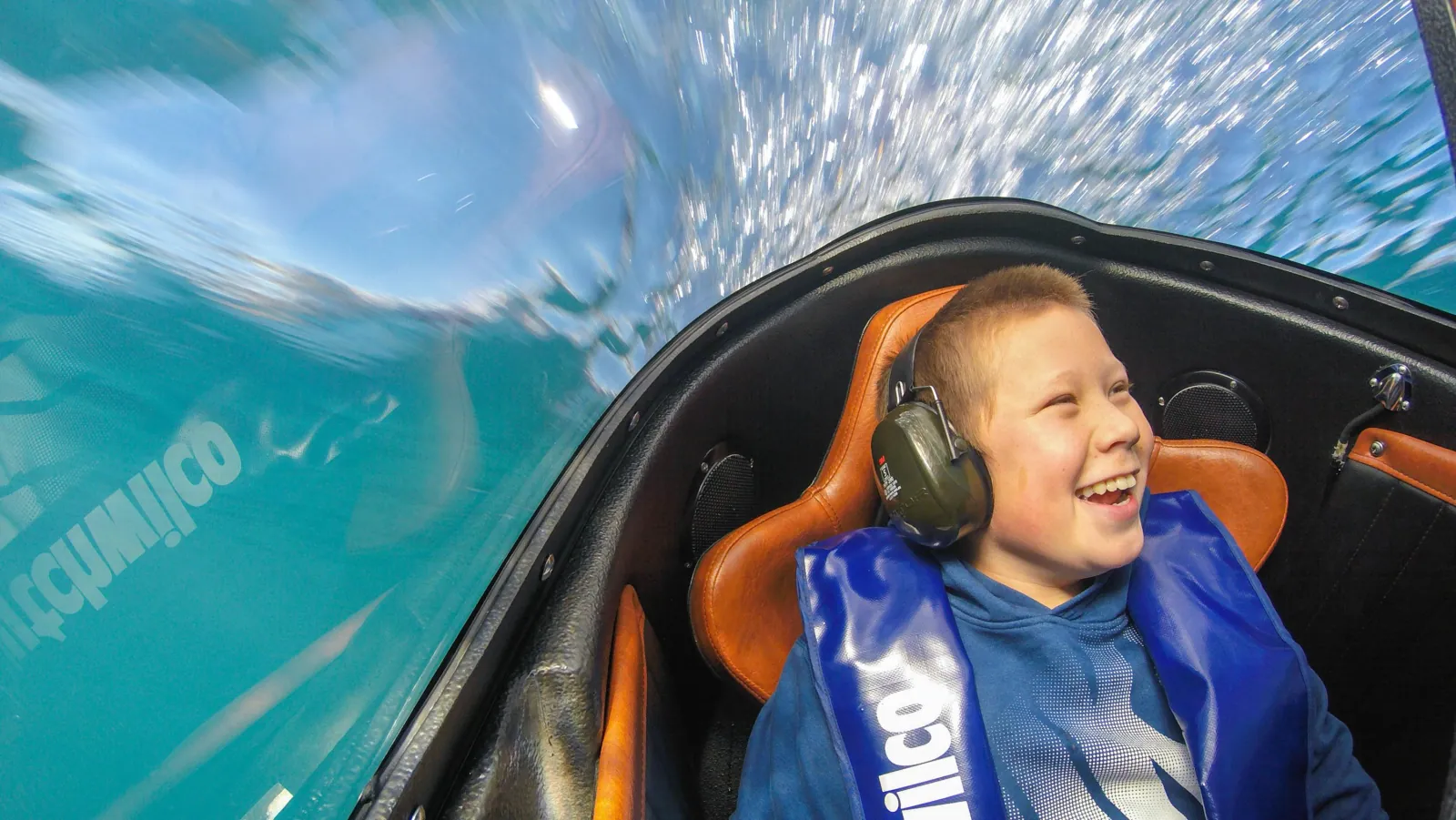 A child grinning inside the sealed cockpit with water washing over the canopy