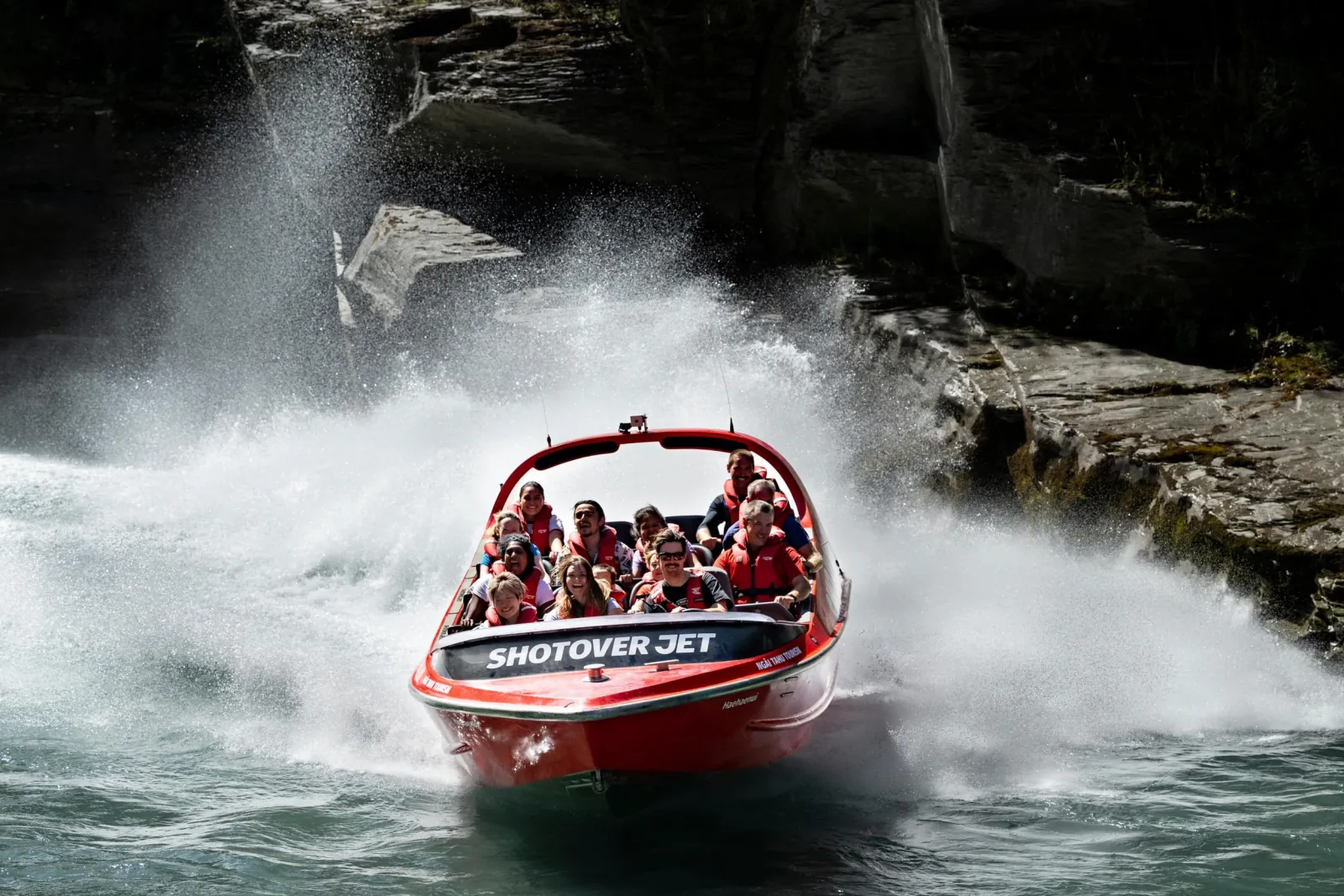 A red Shotover Jet boat throwing a sheet of white spray in a grey rock canyon