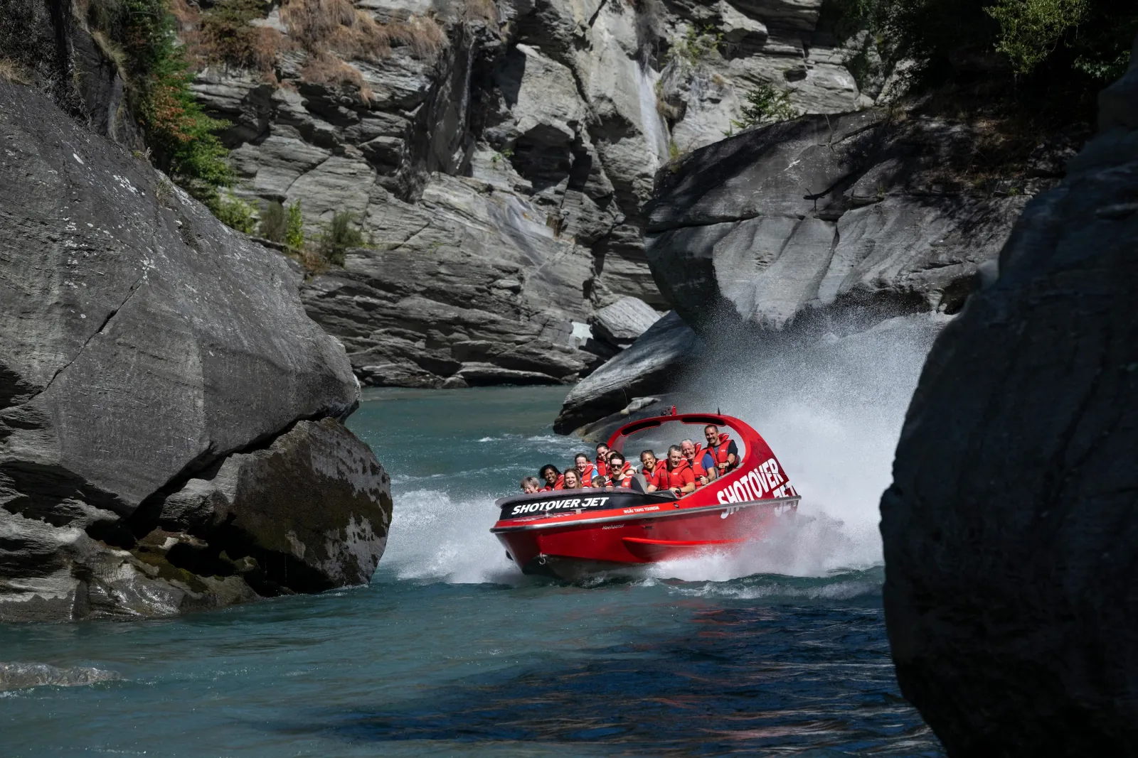 A red jet boat passing close under a dark overhanging rock wall on the Shotover