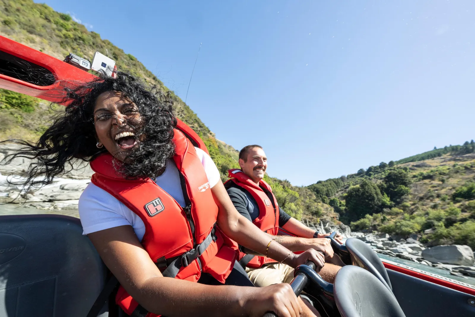 Two passengers in red life jackets laughing aboard a red jet boat in the river valley