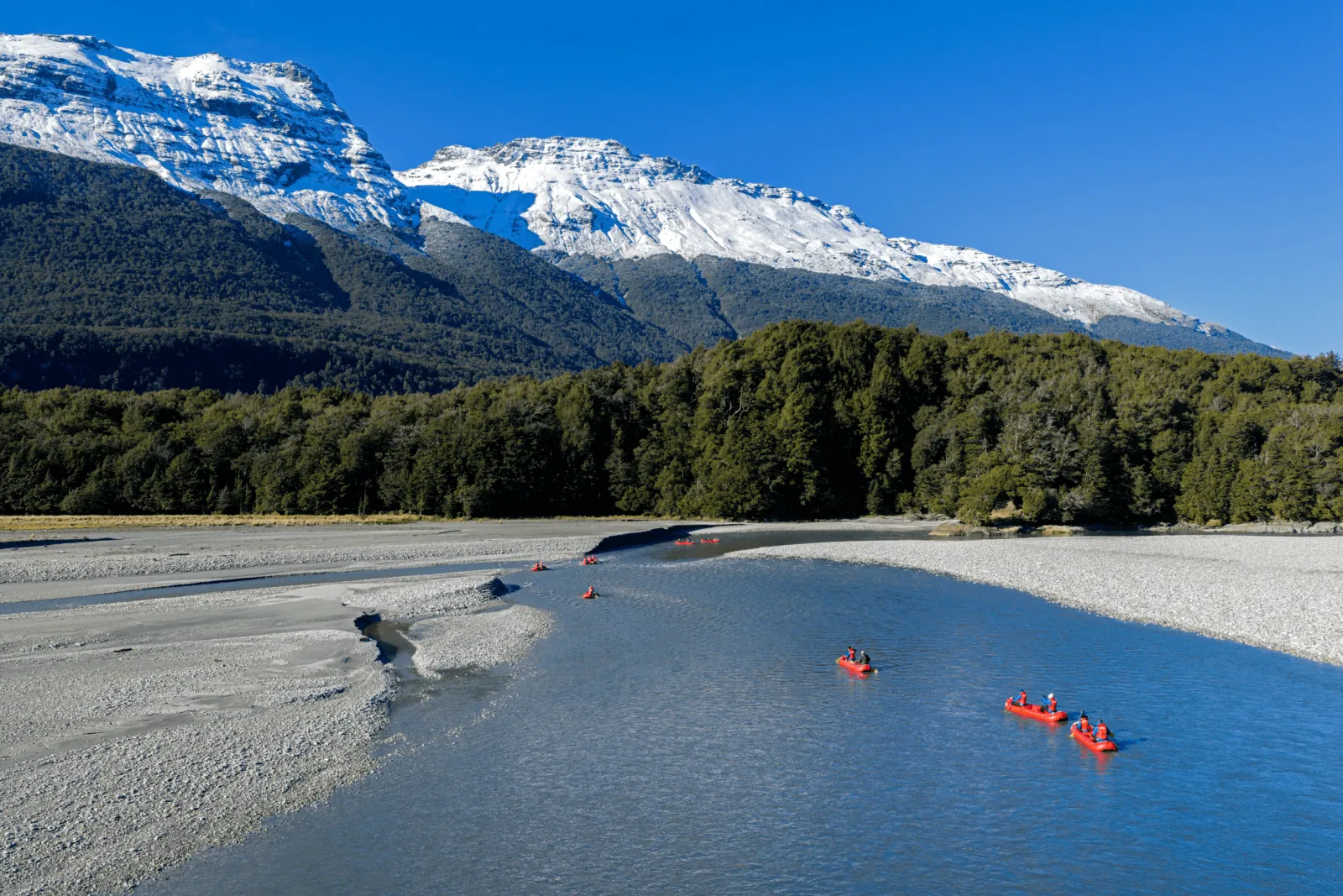 Funyak canoes strung out across a wide flat river under snow-covered peaks