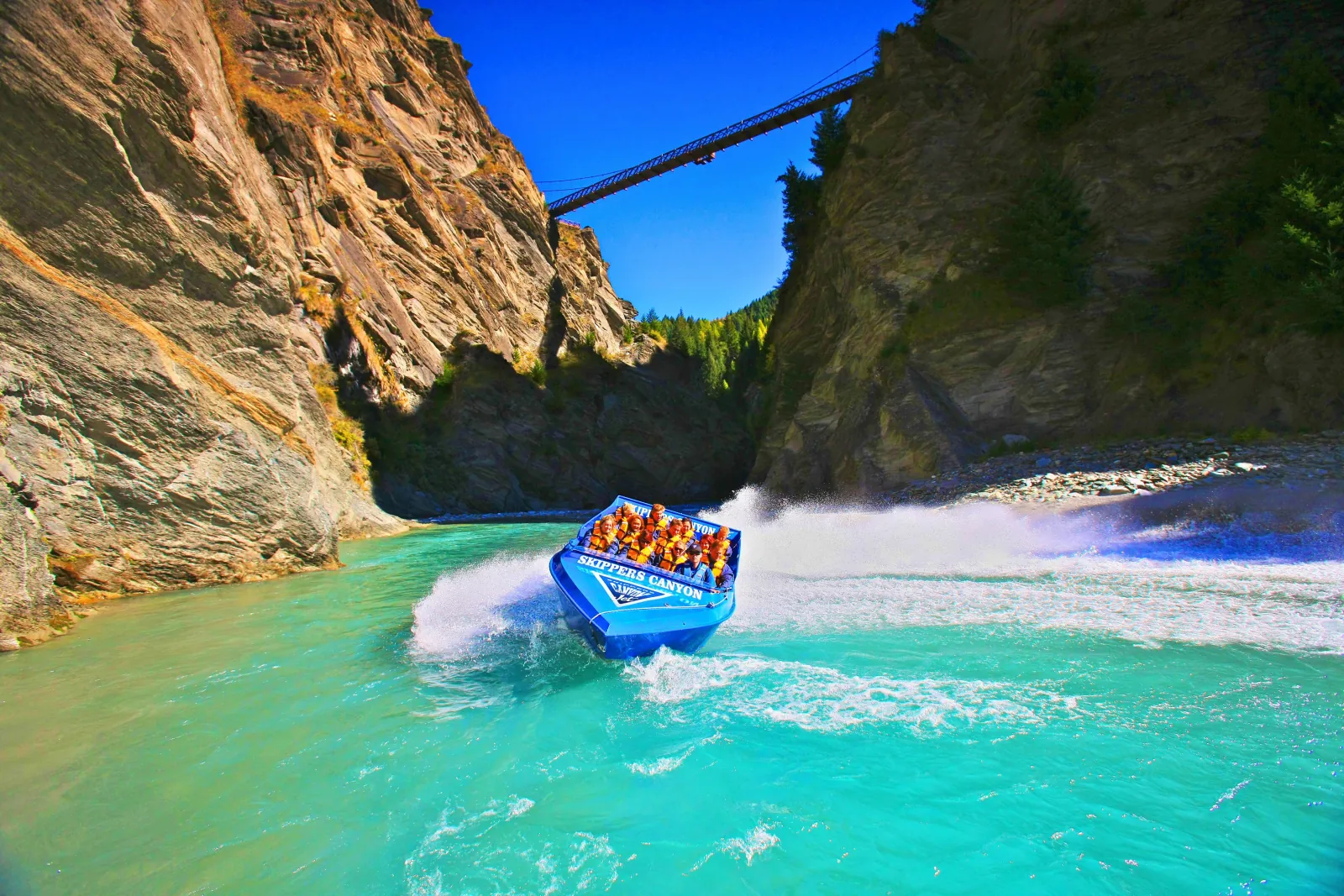 A blue jet boat on turquoise water deep in a narrow canyon, a bridge high above
