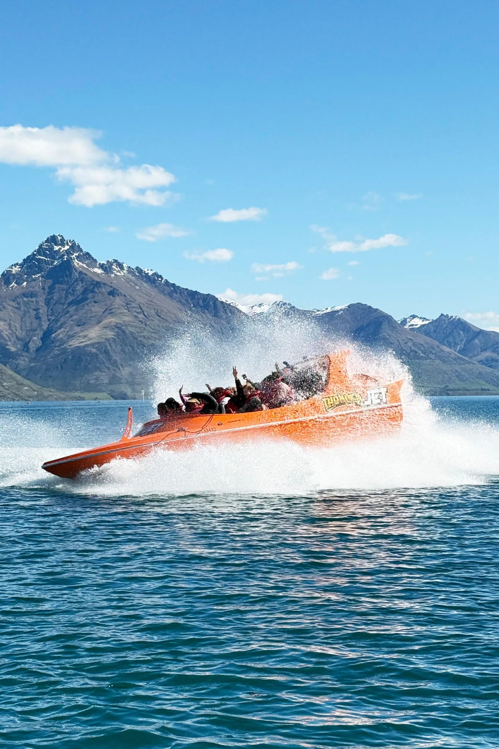 An orange jet boat throwing a wall of spray on open water below the mountains
