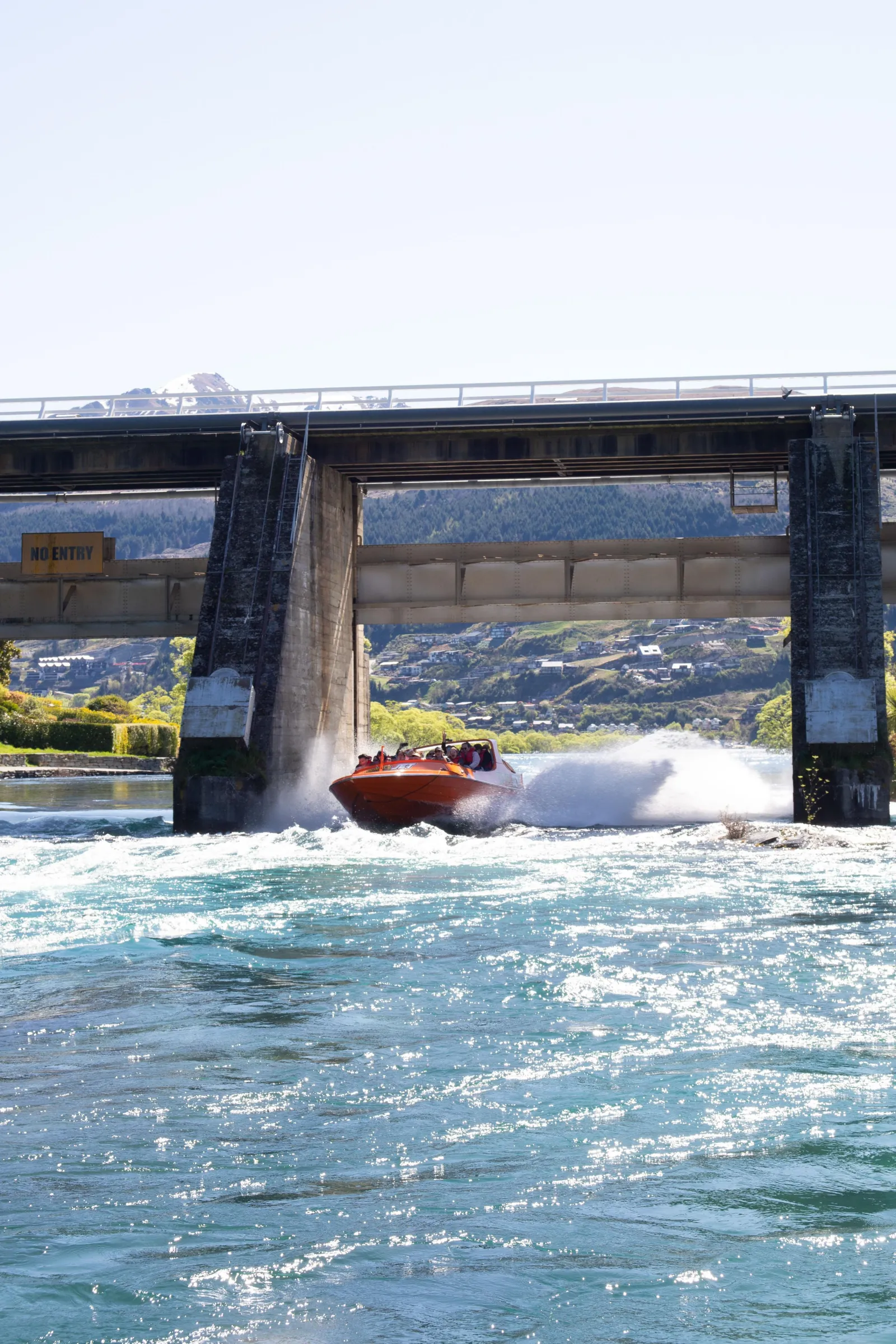 An orange jet boat passing under a low bridge on the river