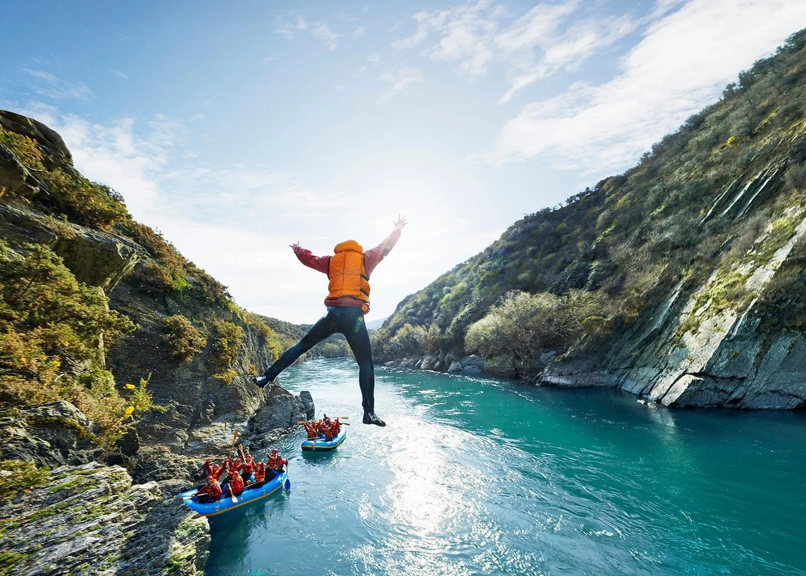 A rafter standing arms-wide on a rock above the river, with rafts on the water below