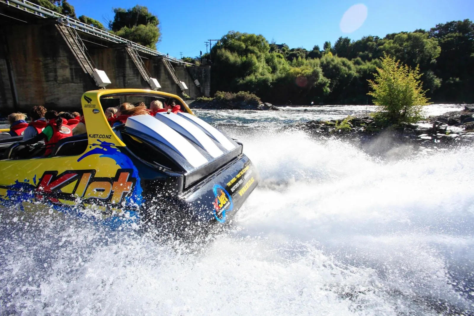 A yellow KJet boat throwing spray as it powers past a low weir