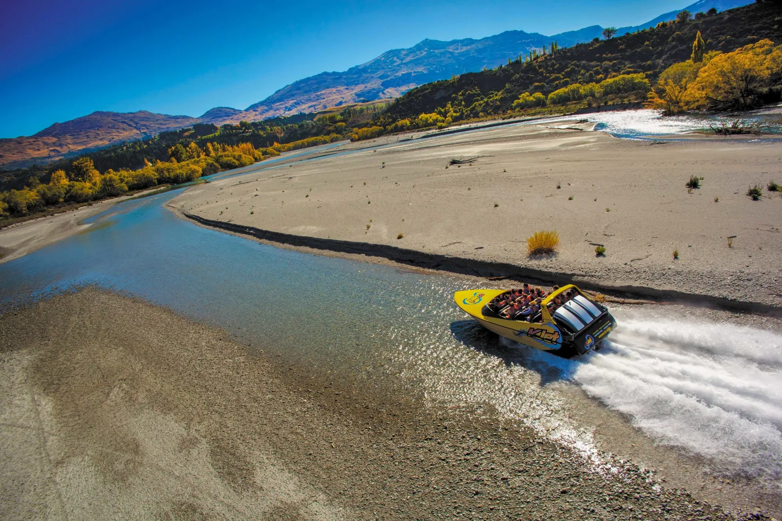 A yellow KJet boat on a wide shingle-lined river bend with autumn trees behind