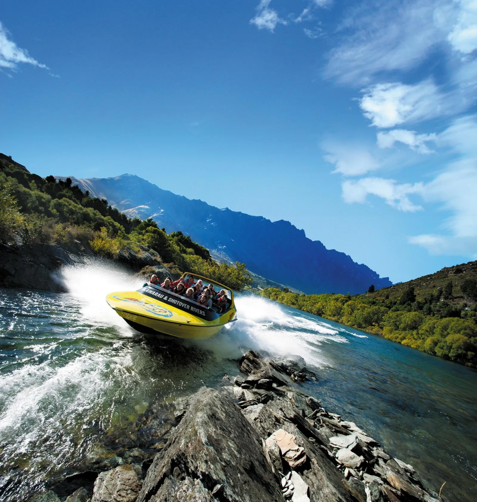 A yellow KJet boat on a shallow reach below tussock hills under a blue sky