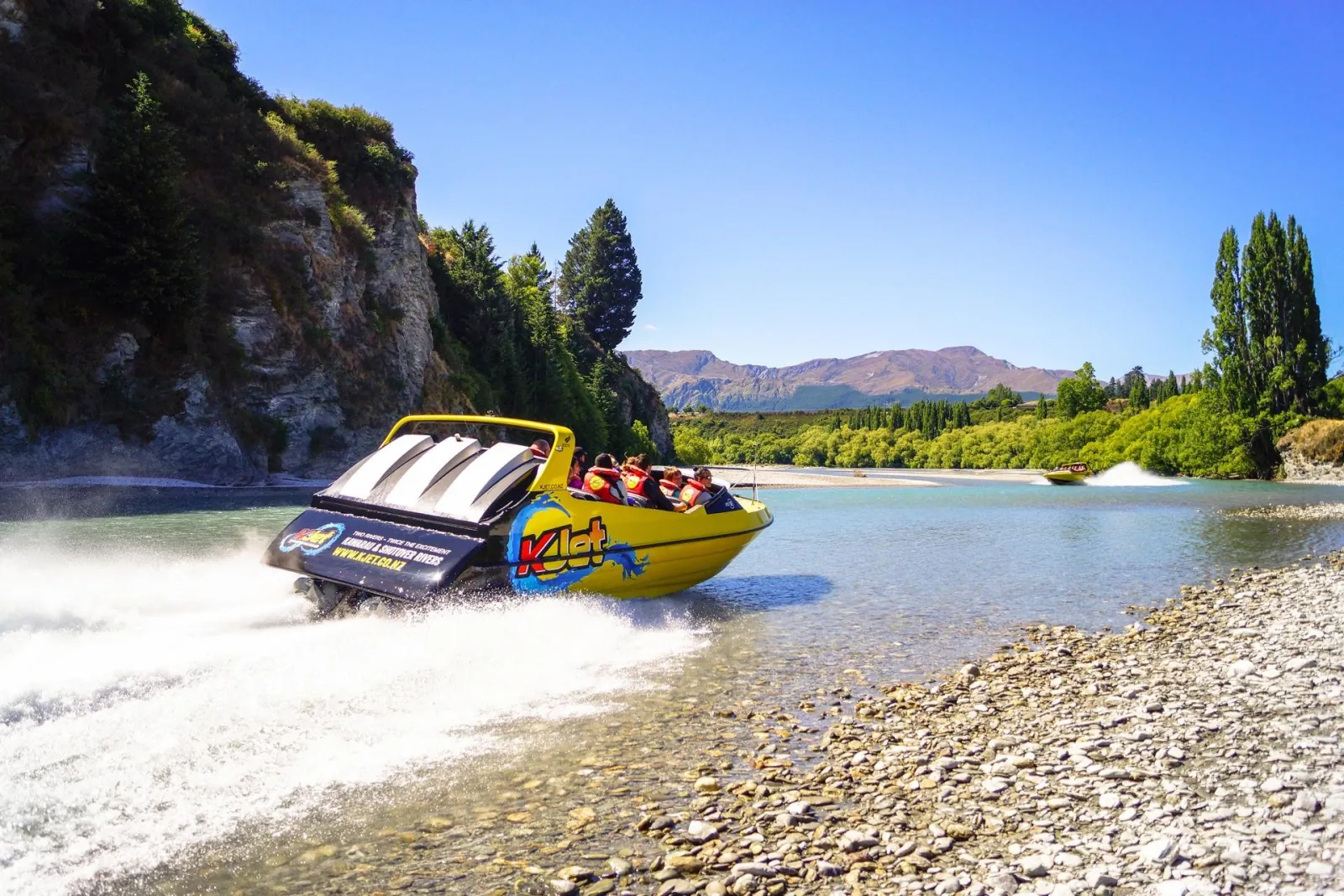 A yellow KJet boat running a shingle channel with mountains behind it
